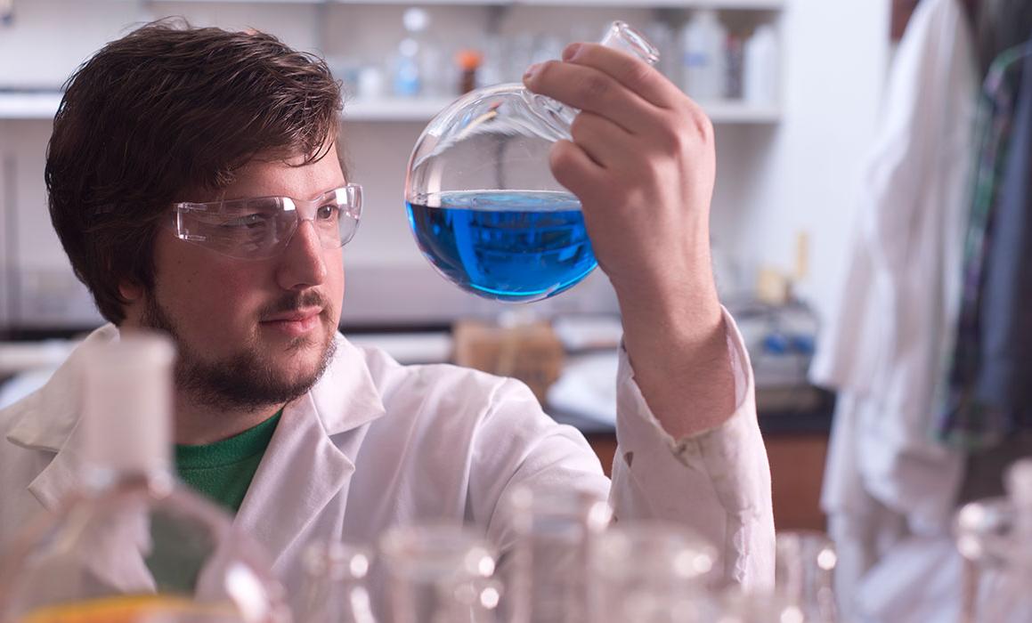 Student presenting as white male in lab coat and safety glasses holds up and looks at round vial with blue liquid in a lab